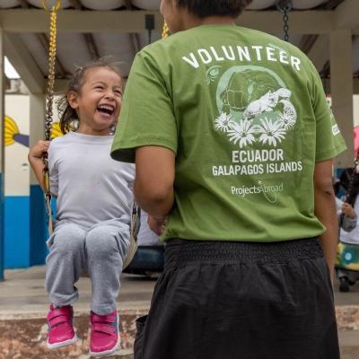 Childcare volunteer plays with a young girl on the swings outside a day care centre in Ecuador. Projects Abroad female volunteer working with children in Ecuador smiles with a child on swing set at our Childcare Project.